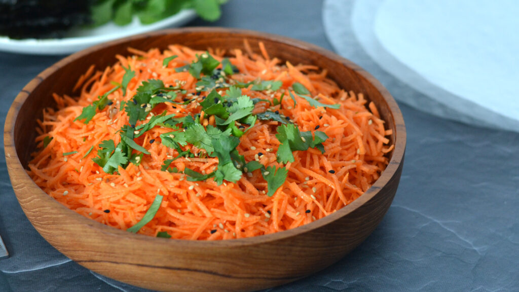 Salade de carottes râpées garnie de sésame et de coriandre dans un bol de bois avec feuille de riz et nori séchés en arrière plan.