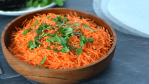 Salade de carottes râpées garnie de sésame et de coriandre dans un bol de bois avec feuille de riz et nori séchés en arrière plan.