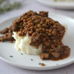 Mijoté de lentilles et champignons Mijoté de lentilles et champignons servi sur une purée de pommes de terre onctueuse, garni de thym dans une assiette en céramique.