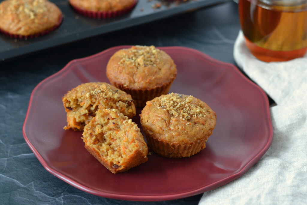 Muffins aux carottes, dattes et thé chaï (ultra moelleux) 3 muffins aux carottes dans une assiette rouge avec un thé chaï en arrière plan. Focus sur un muffin aux carottes coupé en deux montrant les morceaux de dattes et la texture fondante et moelleuse du muffin.