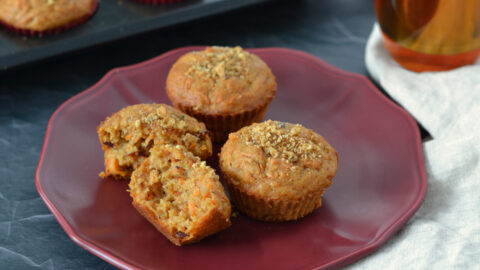3 muffins aux carottes dans une assiette rouge avec un thé chaï en arrière plan. Focus sur un muffin aux carottes coupé en deux montrant les morceaux de dattes et la texture fondante et moelleuse du muffin.