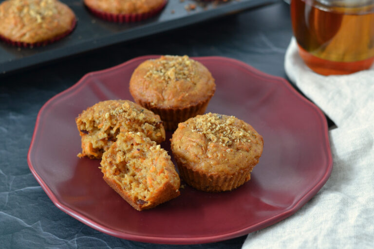 3 muffins aux carottes dans une assiette rouge avec un thé chaï en arrière plan. Focus sur un muffin aux carottes coupé en deux montrant les morceaux de dattes et la texture fondante et moelleuse du muffin.