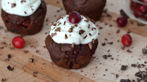 Muffin double chocolat avec sa ganache au yogourt et chocolat blanc couvert de copeaux de chocolat noir râpé avec une canneberges sur le dessus. Deux muffins en arrière plan avec canneberges.