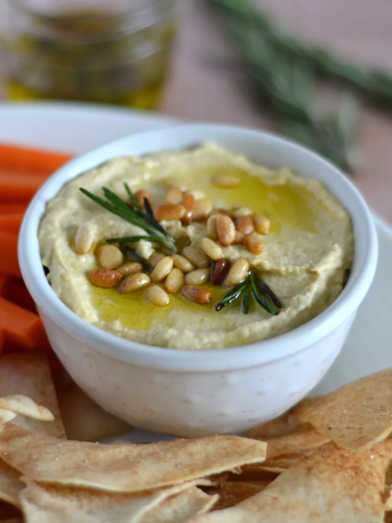 Bol de houmous garni de noix de pin et d'huile d'olive infusée au romarin, dans une assiette de crudités et de chips de pitas