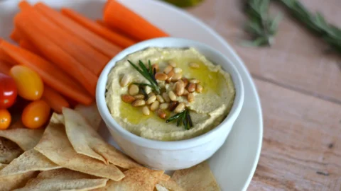 Bol de houmous garni de noix de pin et d'huile d'olive infusée au romarin, dans une assiette de crudités et de chips de pitas