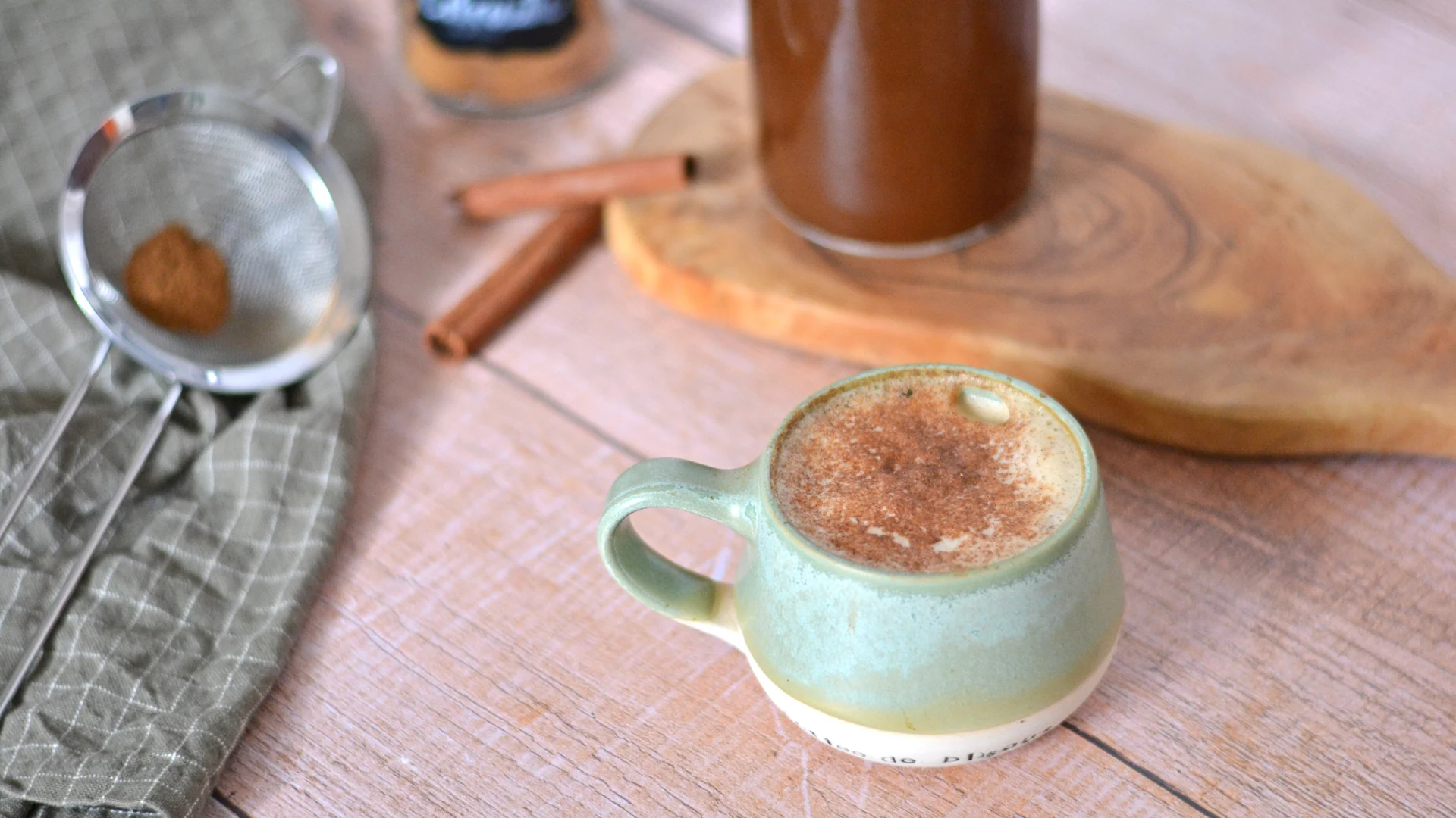 Latté préparé avec du sirop à la citrouille avec en arrière plan, un contenant de sirop, de la poudre d'épices automne et des batônnets de cannelle.